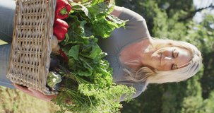 Vertical video of smiling caucasian woman holding basket with fresh vegetables. organic farm and fresh organically grown vegetables.