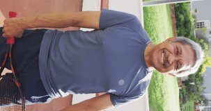 Vertical video of portrait of happy senior biracial man with tennis racket on tennis court. healthy, active retirement.