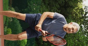 Vertical video of happy senior biracial man playing tennis in garden. healthy and active retirement.