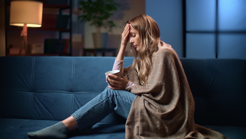 Unwell woman coughing with flu influenza seasonal viral infection disease high temperature chill covered by plaid evening home. Sad female suffering ill sick symptoms epidemic headache pain with tea