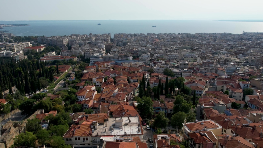 Aerial view of Thessaloniki on a sunny day. White buildings and the fortress drone shot. Flyover rooftops with sea in the background. Travel destination rich with history. Summer day in the city