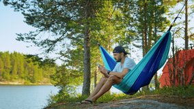Man is sitting in hammock and calling on video on smartphone in camping on lake. go everywhere