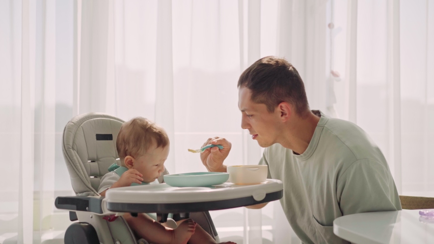 Father acting Mom feeding his son baby on chair in the house. Father