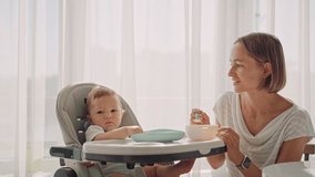Baby sits at a children's little table. Mum feeds the child with porridge. Mother gives baby food from a spoon. High quality 4k footage