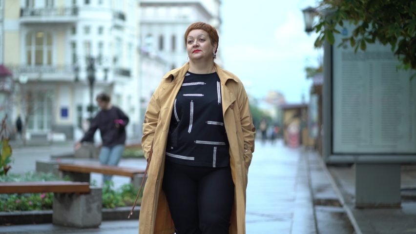 Sad overweight woman in an autumn raincoat walks along the city street. A plump red-haired lady is walking along the sidewalk. Royalty-Free Stock Footage #1094596587