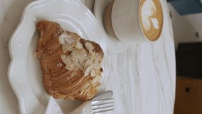 vertical video view of an almond croissant in a plate on the white marble table in cafe with fork and knife utensil in the plate, morning bakery bake meal pastry