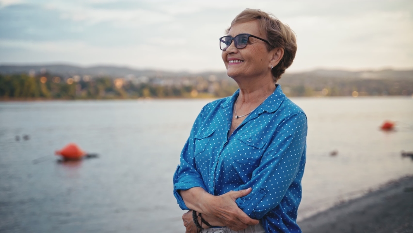 Portrait of a senior caucasian woman taking off her glasses and taking a deep breath while standing on an empty river beach.