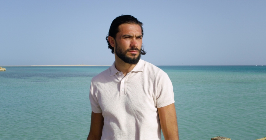 Shot of a man staring at the sea, the man is wearing a white blouse leaving one button open, the man has tanned skin with long black hair, small slow tide waves in the sea behind him
