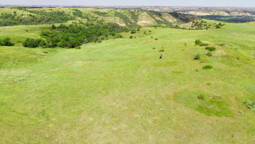 Cowboy riding on horseback in a large open field with rolling hills in the background, aerial flyover