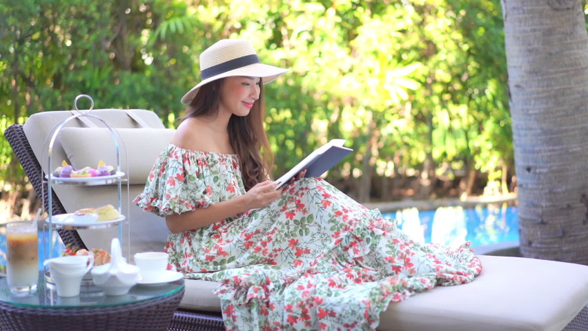 A pretty young woman in a sundress and straw sun hat sits on a chase lounge as she reads her book.  Royalty-Free Stock Footage #1094609731