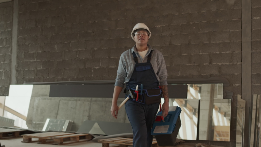Medium long of Biracial woman-construction worker wearing helmet and goggles, carrying toolbox, walking confidently towards camera in building in progress Royalty-Free Stock Footage #1094613199