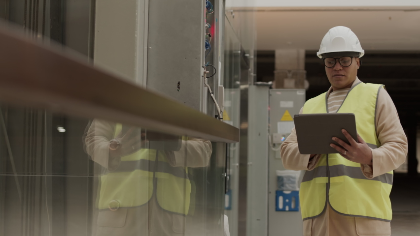 Medium shot of professional Biracial electrician wearing green reflective vest and whit helmet, standing in utility room, holding tablet, conducting diagnostics, then walking away Royalty-Free Stock Footage #1094613829