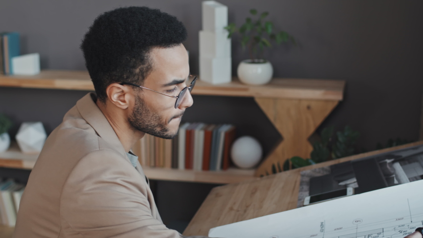 Side-view of young male Biracial architect sitting at desk in office at daytime, drawing blueprint