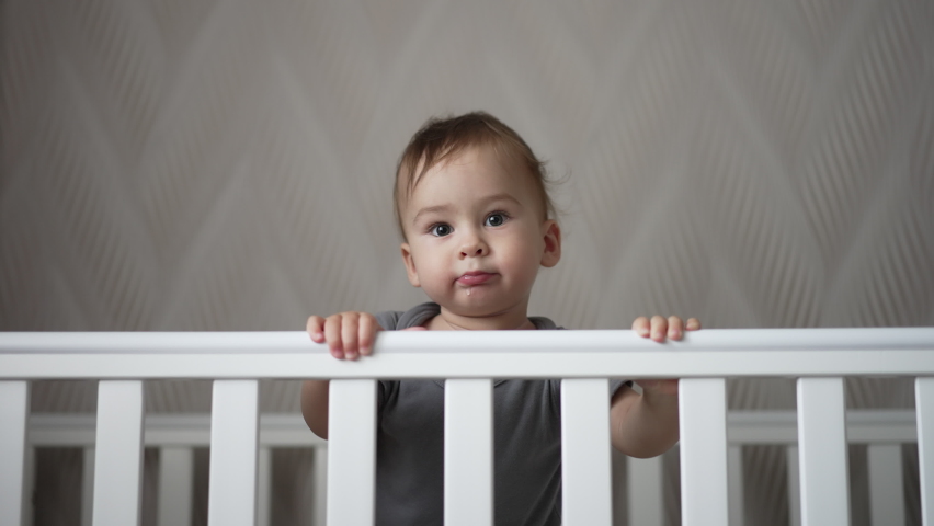 Charming beautiful baby boy standing in his crib. Lovely Caucasian child smiling adorably showing two lower teeth. | Shutterstock HD Video #1094617243