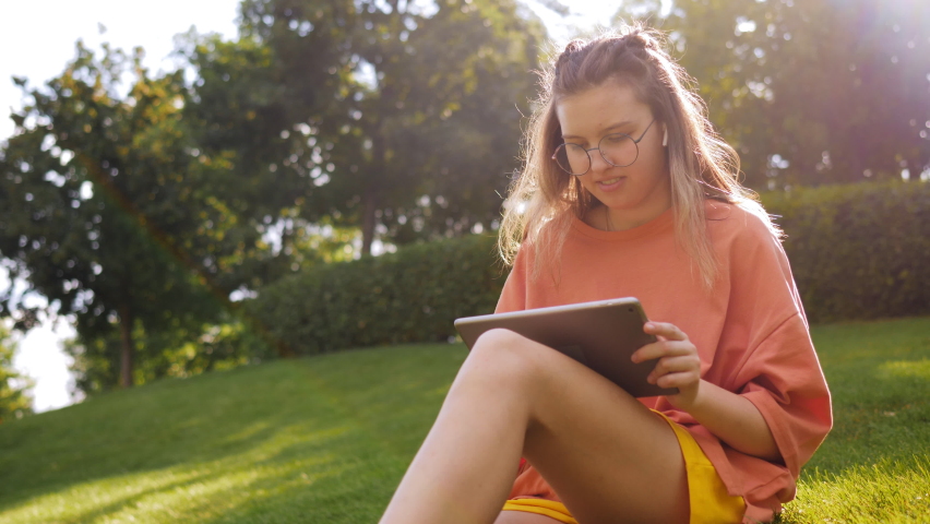 A teenage girl on a green lawn in the park is sitting with a tablet in her hands doing her studies.