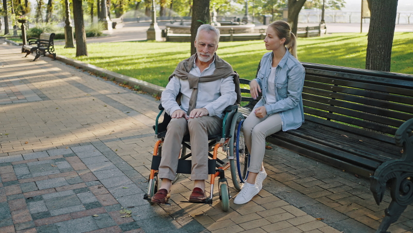 Woman hugs elderly bearded father in wheelchair sitting on wooden bench after walk in park.  pensioner enjoys time with daughter in sunny morning