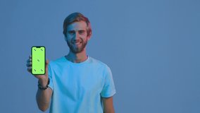 Smiling modern young guy walks, showing smartphone with mockup blank green screen, advertising mobile apps. Copy space