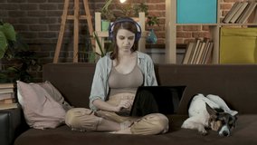 Attractive girl with headphones is sitting on cozy sofa at home in living room using laptop for study or work. high school student uses laptop to search for information on Internet and communicate