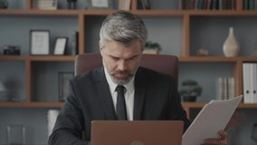 Middle-aged bearded businessman sitting in his office studying profit charts and comparing them with digital statistics