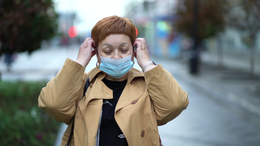 An elegant woman with short red hair walks around the city and puts on a medical mask on her face. Woman protecting herself from the virus with a mask. | Shutterstock HD Video #1094632709