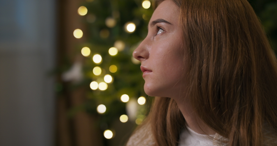 Close-up of sad frustrated girl sitting by Christmas tree holding cup of tea. She has long nails covered with black varnish. The girl broke up with boyfriend. She almost cries.