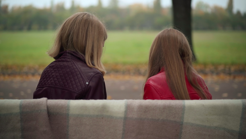 Back view teen friends sitting on bench in park turning looking at camera. Positive cheerful Caucasian teenage girls in coronavirus face masks posing outdoors on autumn weekend | Shutterstock HD Video #1094635383
