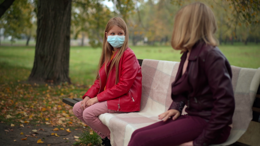 Cute girl in Covid face mask stretching hands with friend sitting on bench maintaining social distance. Positive Caucasian teenagers meeting in autumn park on coronavirus pandemic | Shutterstock HD Video #1094635401