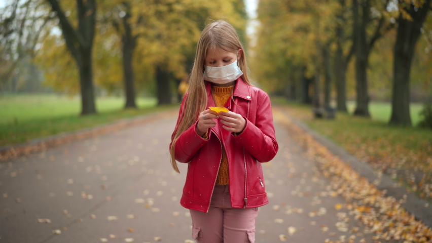Depressed teen girl in Covid face mask standing on alley in park playing with golden leaf. Medium shot portrait of frustrated Caucasian teenager alone outdoors on overcast cloudy autumn day | Shutterstock HD Video #1094635447