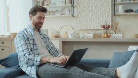 A smiling Caucasian man is working on a laptop while sitting on the couch in his cozy smart apartment. Happy guy working remotely with enthusiasm. Browsing the Internet, working from home