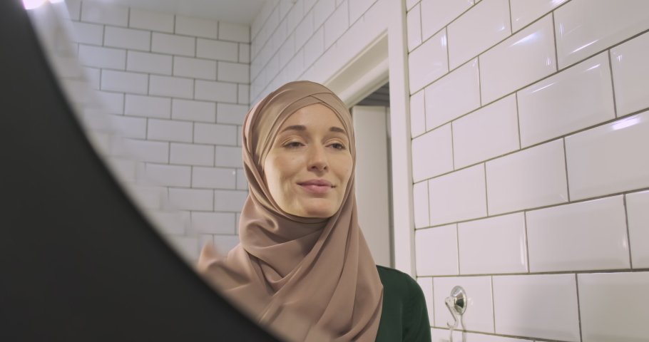 Reflection of a Muslim woman in the mirror. Happy girl in hijab applies make-up with a brush. Concept of a modern woman, independence, female beauty. | Shutterstock HD Video #1094649251