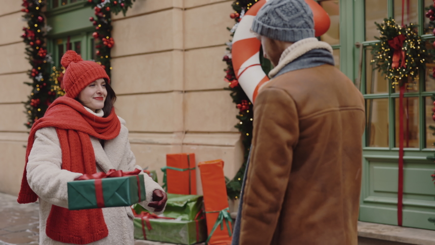 The lovely couple is hugging in the street giving each other christmas gifts and feeling happy and in love. Back view of the man hugging his girlfriend and she is holding the christmas gift