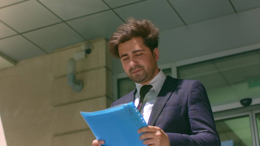 Portrait of successful businessman wearing stylish suit holding documents walking outdoor near the office buildings.