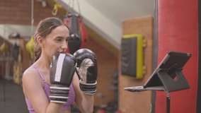 a girl in a pink top beats a punching bag in black boxing gloves. teaches by video link how to do the exercise correctly