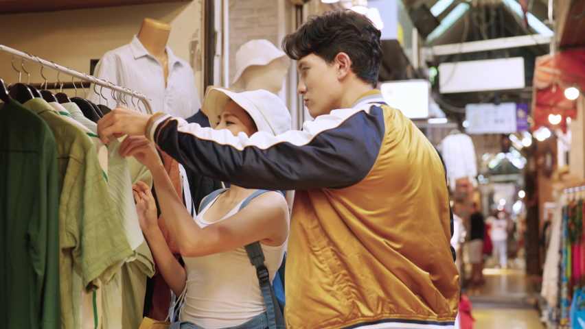 4K Young Asian couple enjoy and fun outdoor lifestyle shopping together at street market on summer holiday vacation. Happy man and woman choosing and buying fashion clothes together at clothing shop.
