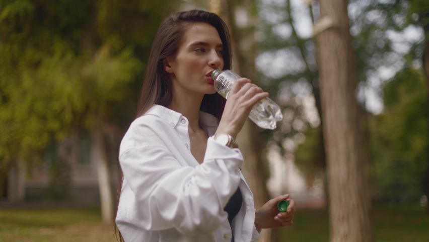 A young woman drinking water from a bottle while walking in the park taking a break to drink water. Young woman long haired brunette drinking water outdoors from bottle | Shutterstock HD Video #1094666893