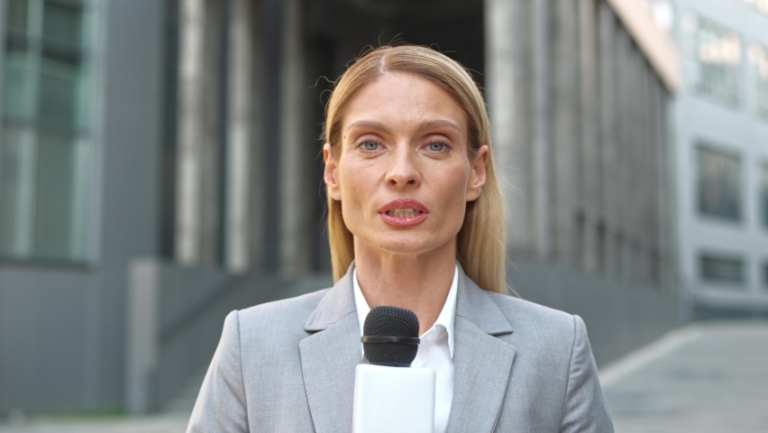Close up of young beautiful female anchor reporting directly from street in front of administrative office building. Woman TV correspondent broadcasting live talking in microphone. News concept