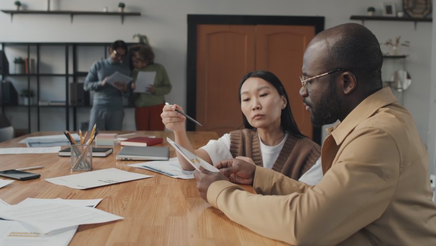 Medium side view of young African American man and Asian woman sitting at table in conference room, having conversation, working with documents