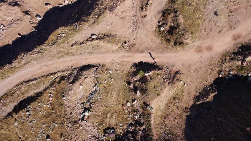 A man walks along a dirt road among yellow hills. There are green bushes and grass in places, but everything else is yellow. Large stones lie on the sides. The long shadow of a man and ravines.