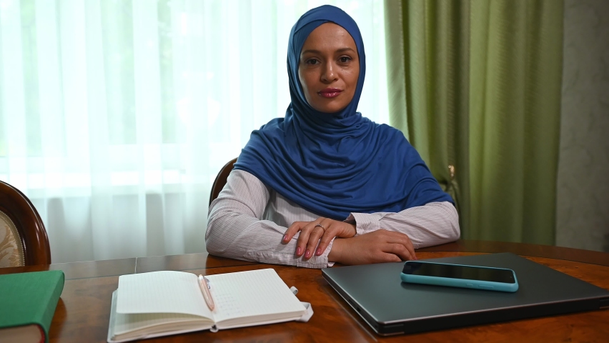 Confident portrait of charming Muslim woman of Middle-Eastern ethnicity, wearing blue hijab confidently looking at the camera, sitting at a wooden table with laptop and smartphone in the meeting room | Shutterstock HD Video #1094675763