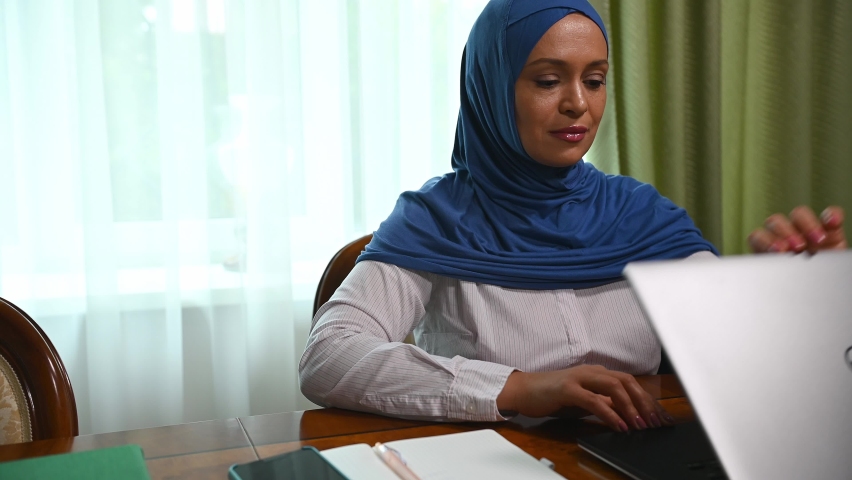 Stylish confident modern Arabic businesswoman, wearing casual shirt and blue hijab, successful financial manager opens the lid of laptop computer. Online business. Work opportunities for Muslim women | Shutterstock HD Video #1094675767
