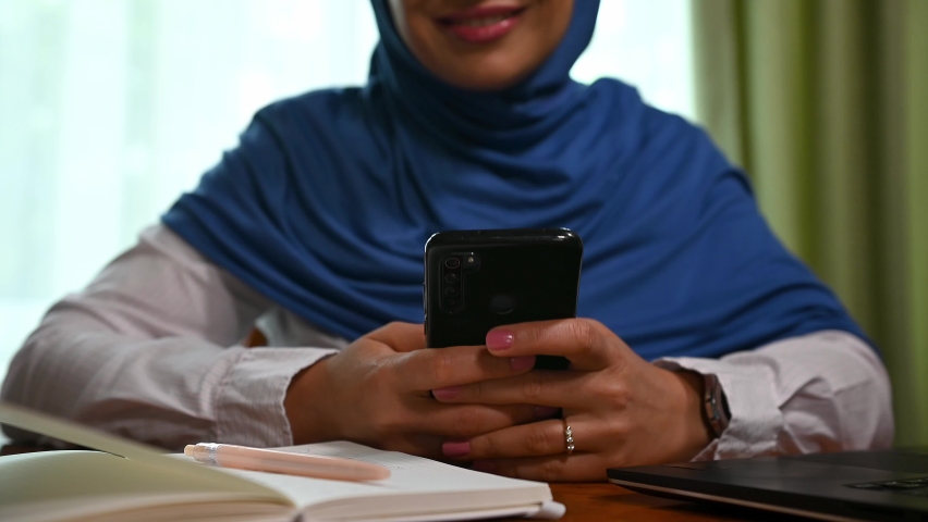 Selective focus on the hands of a pretty Muslim woman of Middle-Eastern ethnicity in blue hijab using smartphone, texting message, checking news feed, browsing websites. Online communication. | Shutterstock HD Video #1094675769