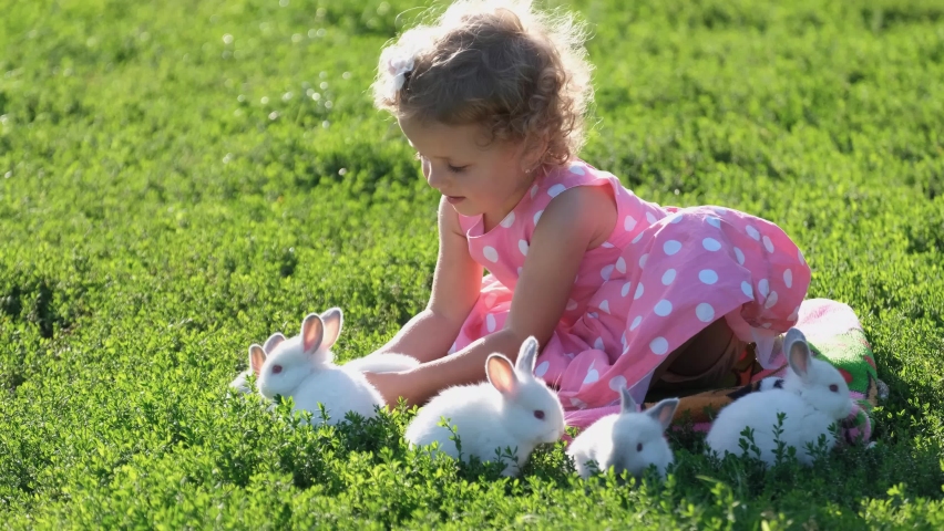 A child plays with white rabbits on the green grass in the garden. Happy child with pets.