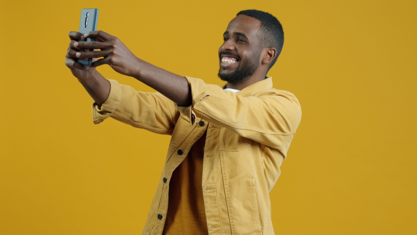 Portrait of handsome Afican American man taking selfie with mobile camera posing on yellow background. Photography and modern technology concept. | Shutterstock HD Video #1094684059