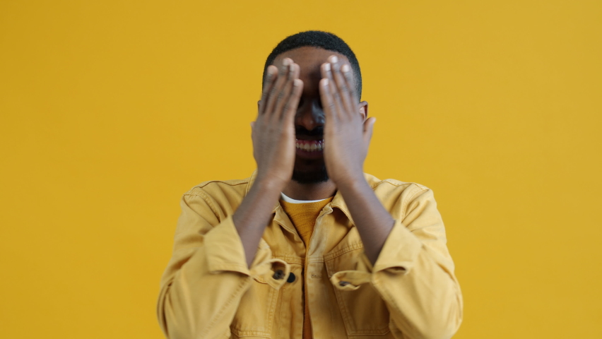 Slow motion of joyful Afican American man covering and uncovering face with hands and making funny grimaces having fun playing hide-and-seek on yellow background | Shutterstock HD Video #1094684061