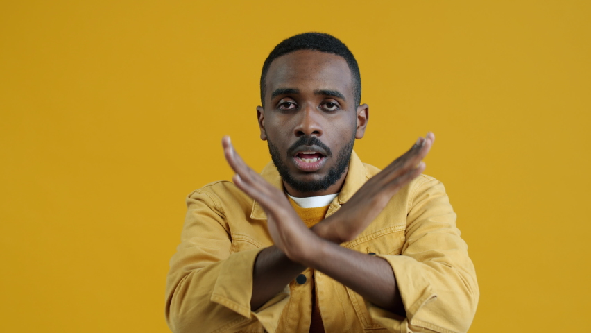 Portrait of serious Afican American man stretching arm with No hand gesture shaking head and looking at camera on yellow color background. People and expressions concept. | Shutterstock HD Video #1094684071