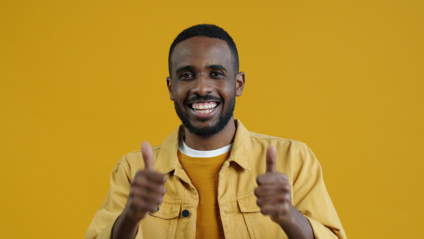 Portrait of excited Afican American man showing thumbs-up hand gesture and looking at camera with happy smile on yellow color background | Shutterstock HD Video #1094684083