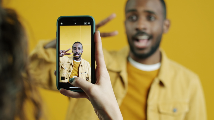 Playful Afican American guy is posing for smartphone camera while hand is taking pictures touching screen on yellow background. Focus is on gadget display. | Shutterstock HD Video #1094684085