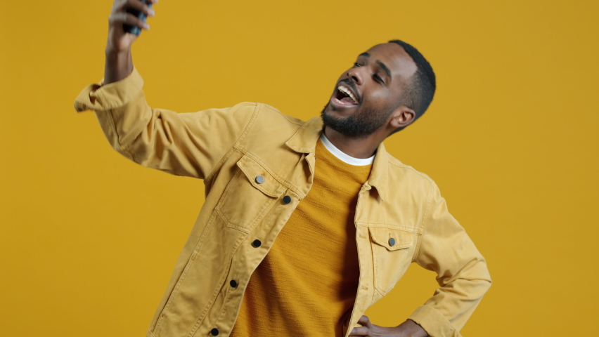 Stylish Afican American student taking selfie with sartphone camera posing and smiling against yellow color background. People and activities concept. | Shutterstock HD Video #1094684091