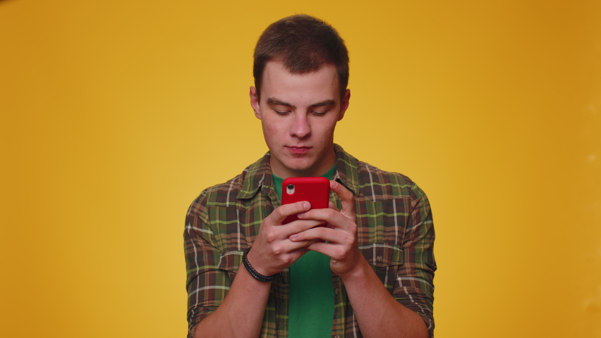 Teen man in shirt use mobile cell phone typing browsing, loses becoming surprised sudden lottery results, bad fortune, loss, fail. Young adult guy boy isolated alone on yellow studio wall background | Shutterstock HD Video #1094687461