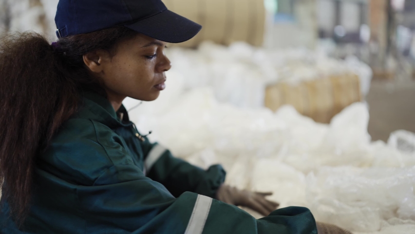 An African-American woman in a special uniform sorts polyethylene at a waste recycling plant. Processing of raw materials, recycling. Pollution control | Shutterstock HD Video #1094690247